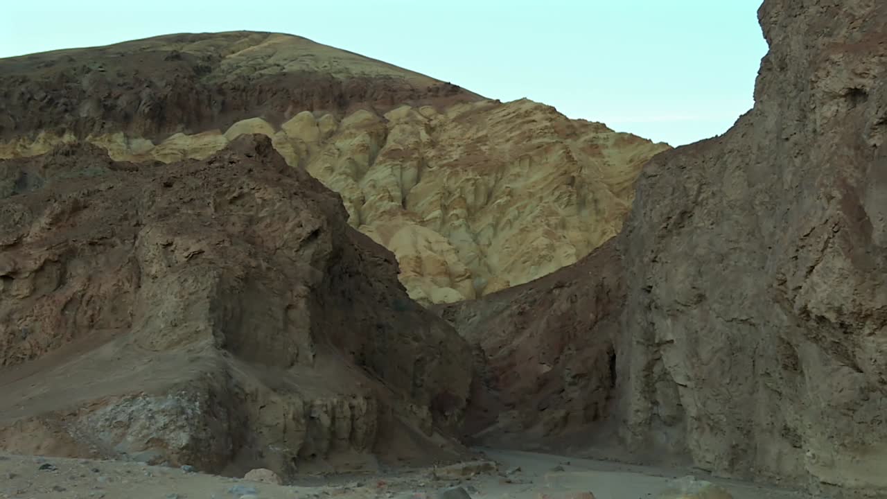 Layered eroded canyon walls with pale yellow and red rock formations framed by steep cliffs in Death Valley National Park, California, USA. Zoom out view