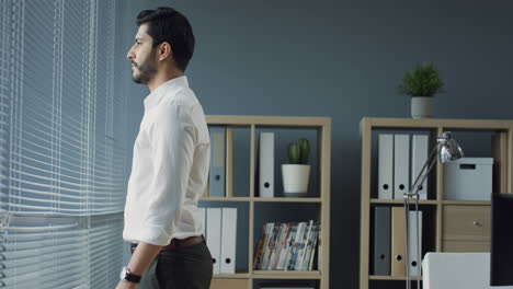 arabian businessman standing in his cozy modern office and looking out the window, then he turns to the camera, crosses his arms and smiles cheerfully