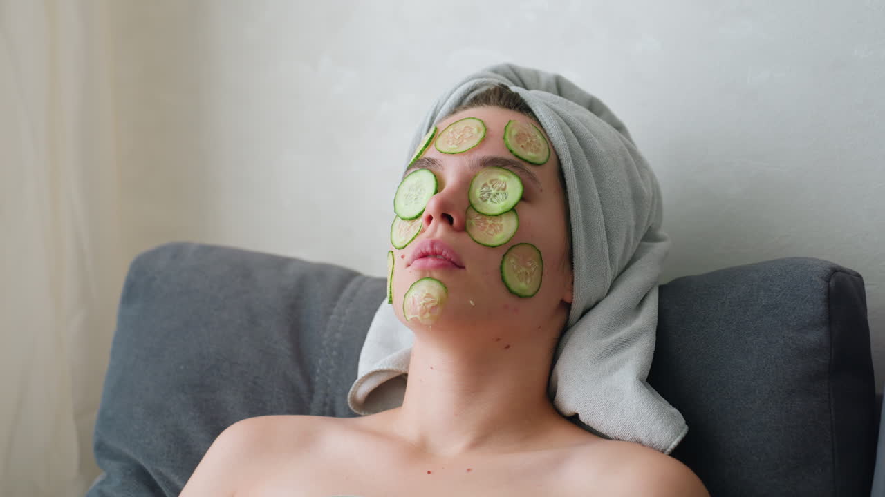 Close-up of woman with hair wrapped in towel, cucumber slices placed on her face, relaxing in a peaceful self-care routine, she enjoys a soothing facial treatment while unwinding at home