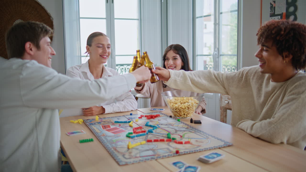 Friends clinking beer bottles at wooden table closeup. People playing board game