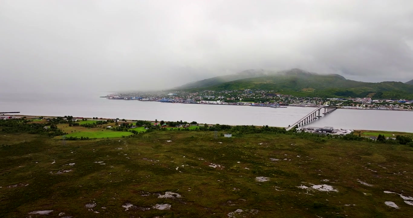 Weite Luftaufnahme mit Drohne von Sortland in Vesterålen, Nordnorwegen. Blick auf die Brücke über dem Wasser mit nebelverhangenem Berg und bewölktem Himmel im Hintergrund.