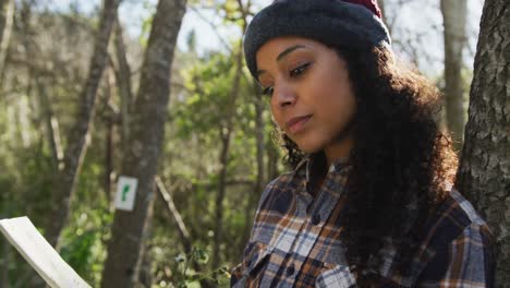 biracial woman checking map in forest during hiking in countryside