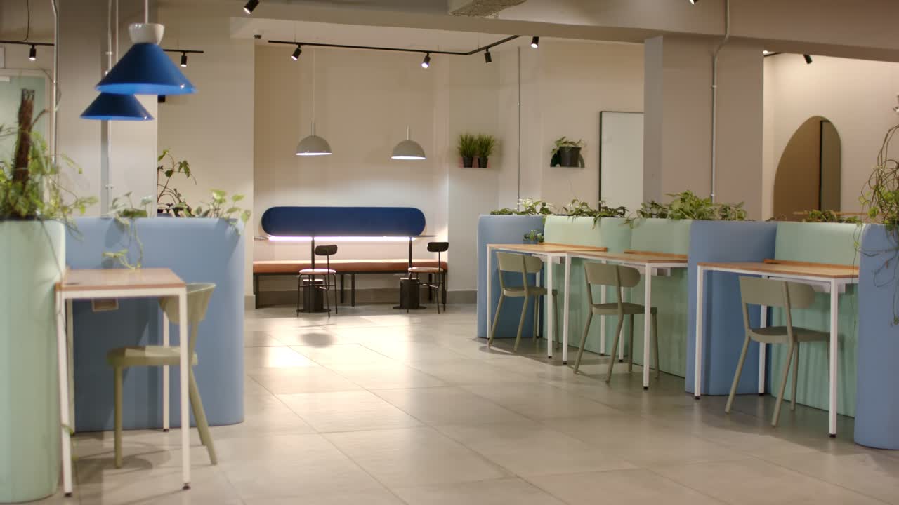 La cámara panea hacia la derecha mostrando lámparas y plantas en una sala de coworking, revelando zonas de asiento.