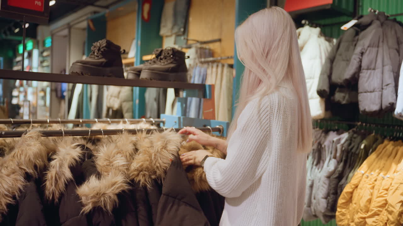 Urban shopper moves to winter coat rack browsing fur trimmed coats and shoes displayed above feeling fabric quality and exploring style under ambient store lighting amid apparel racks