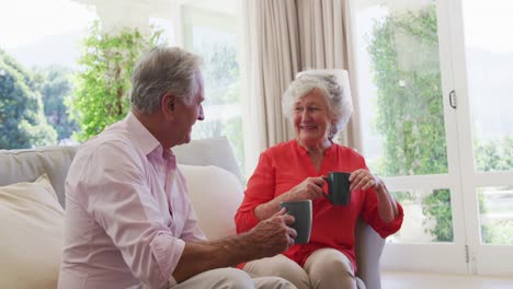 Glückliches-Kaukasisches-älteres-Paar,-Das-Im-Sonnigen-Wohnzimmer-Sitzt-Und-Sich-Unterhält-Und-Eine-Tasse-Kaffee-Trinkt