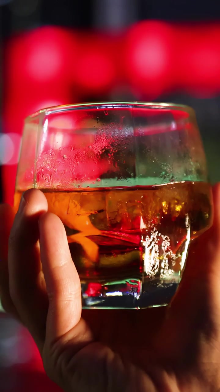 Una mano ruota lentamente un bicchiere di whisky con ghiaccio su uno sfondo di bar vivido e colorato. L'illuminazione calda evidenzia la condensa e i riflessi sul bicchiere.