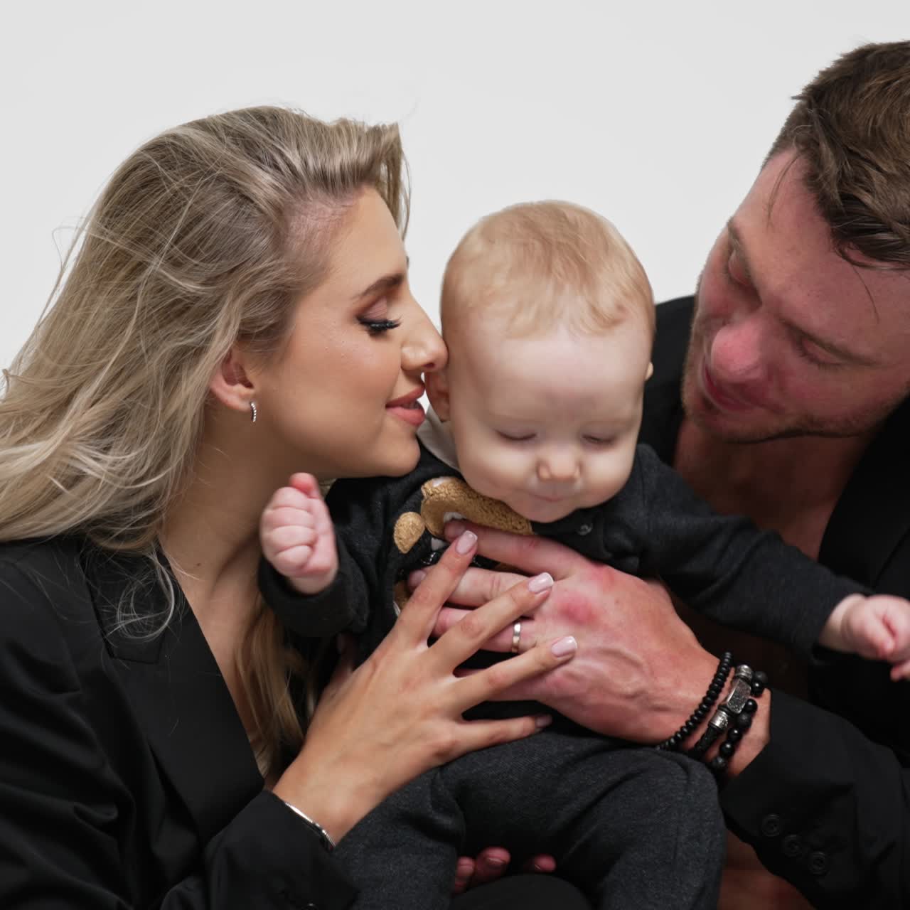 Hübsches kaukasisches Paar mit schwarzen Jacken hält sein süßes blondes Baby. Eltern genießen Elternschaft. Weißer Studiohintergrund
