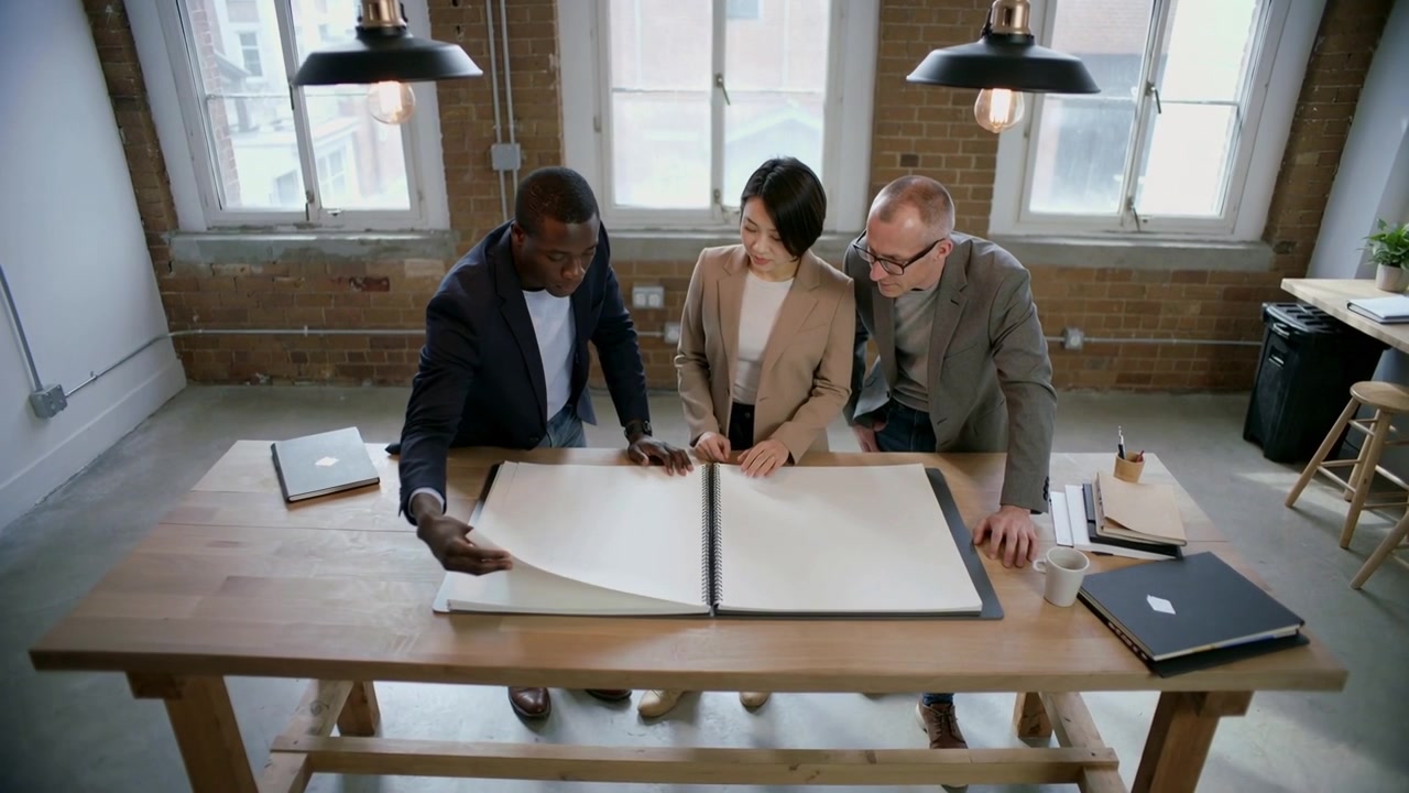 Reaching left man starting turning pages, three team members in blazers reviewing folio in loft