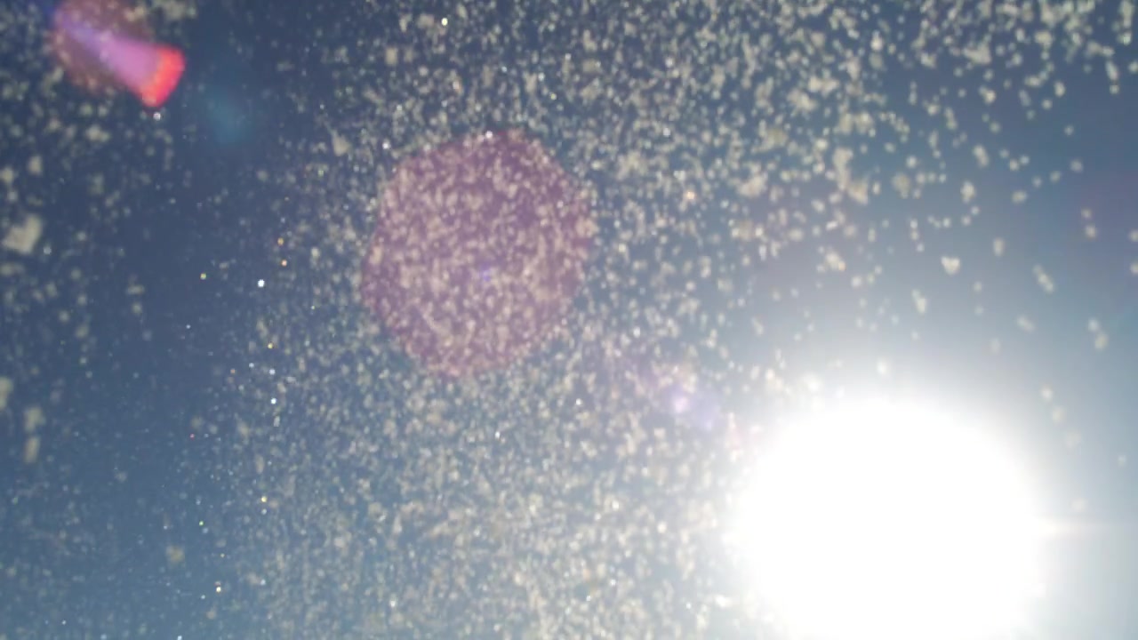 Fine snow spray sparkles around bright backlit sun against blue sky in slow motion
