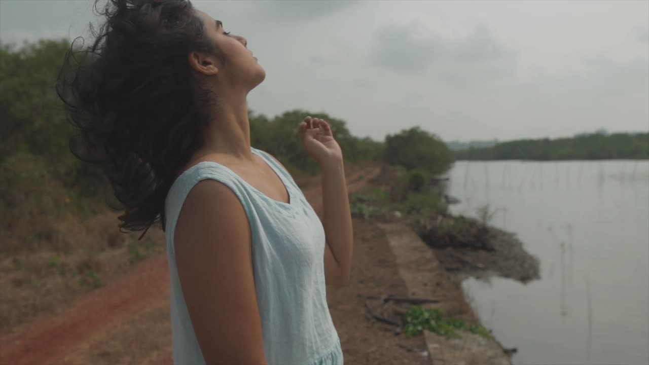 toma manual en cámara lenta de una mujer india joven y elegante con un vestido turquesa acariciando su cabello y mirando a la cámara con una mirada sexy frente a un lago en la naturaleza