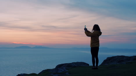 mujer tomando una foto de la puesta de sol sobre el océano desde un acantilado