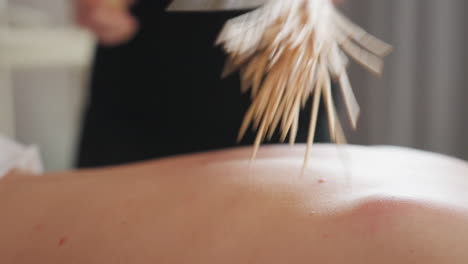 extreme close-up of bamboo massage broom pressing against fair skin woman s back during soothing wellness treatment, emphasizing gentle contact, wood texture, and calming environment in soft focus