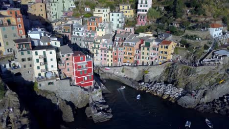 Town-of-Riomaggiore-in-the-Cinque-Terre-Italian-coast-with-boats-and-people,-Aerial-pan-right-shot