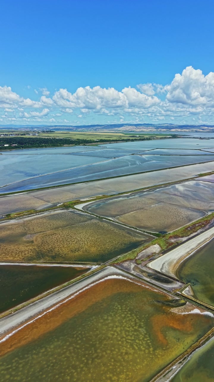 Vue de marais salants et de plans d'eau sous un ciel bleu et des nuages, en journée, dans une zone côtière.