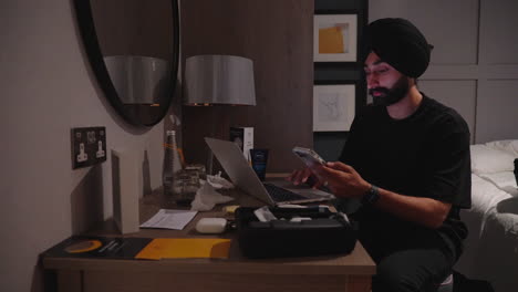 Indian-Man-Sitting-On-His-Working-Table-Using-Cell-Phone-And-Laptop-Inside-The-Bedroom