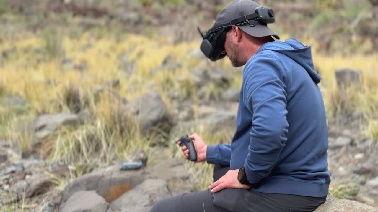Man with FPV goggles and joystick controller operating a quadcopter in a rocky, natural environment
