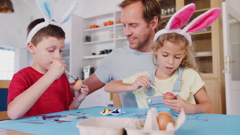 father with children wearing rabbit ears decorating easter eggs at home together