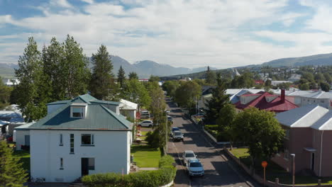 Forwards-fly-above-street-lined-with-houses-in-calm-residential-town-borough.-Mountain-ridge-in-background.-Akureyri,-Iceland
