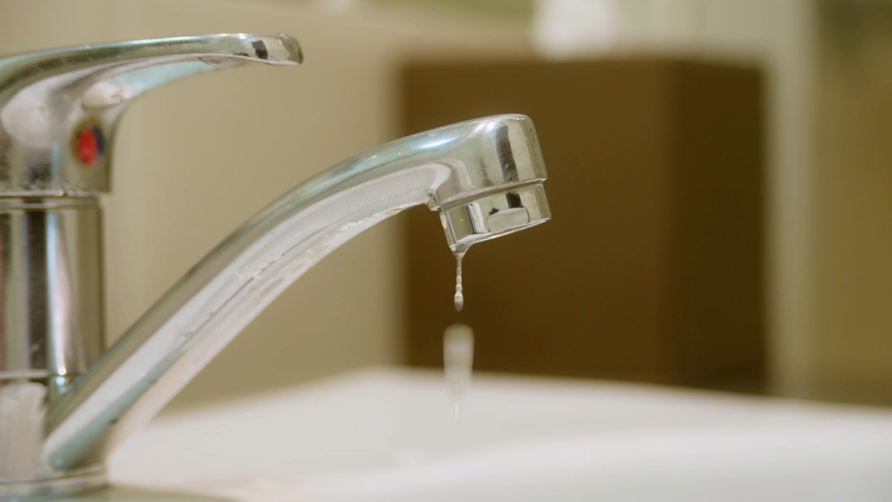 Close up of a bathroom faucet tap slowly dripping water, symbolising water waste, leaks and household utility cost. Plumbing, conservation and environmental concept. Clip 1