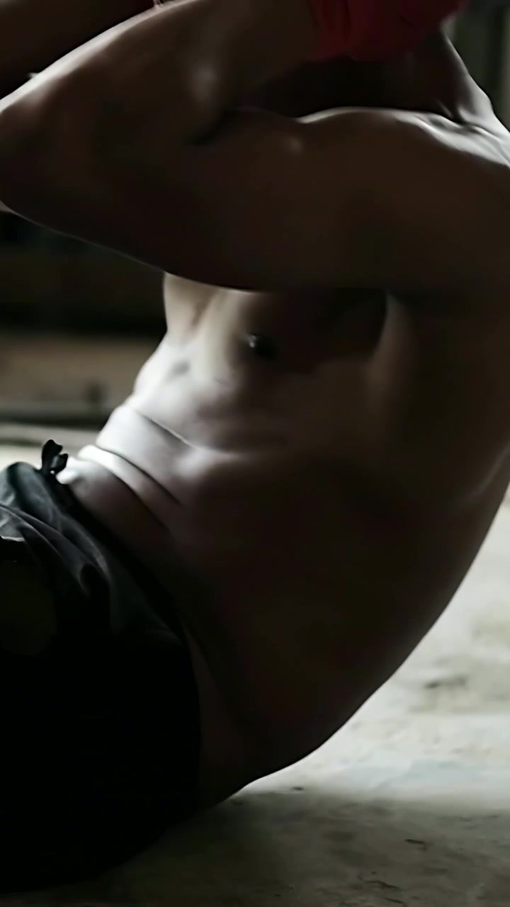 Video verticale: Pugile a torso nudo che inizia a fare addominali in un magazzino, con pantaloncini neri e fasce rosse.