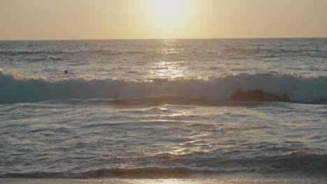 large wave from the pacific ocean bursts onto the shallows of a sandy beach in mexico as several individuals swim during sunset