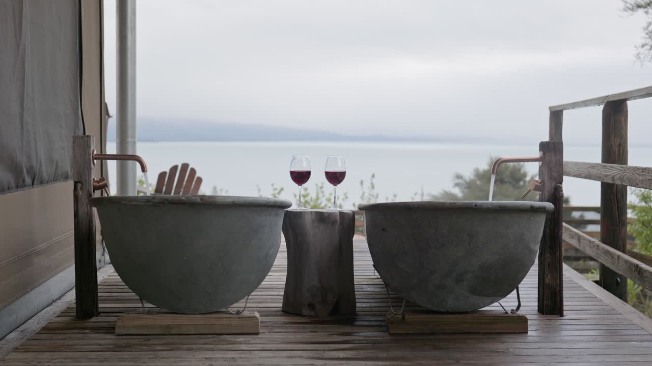 Empty outdoor metal bathtubs on wooden deck with wine glasses and ocean view under overcast sky in luxury New Zealand spa setting