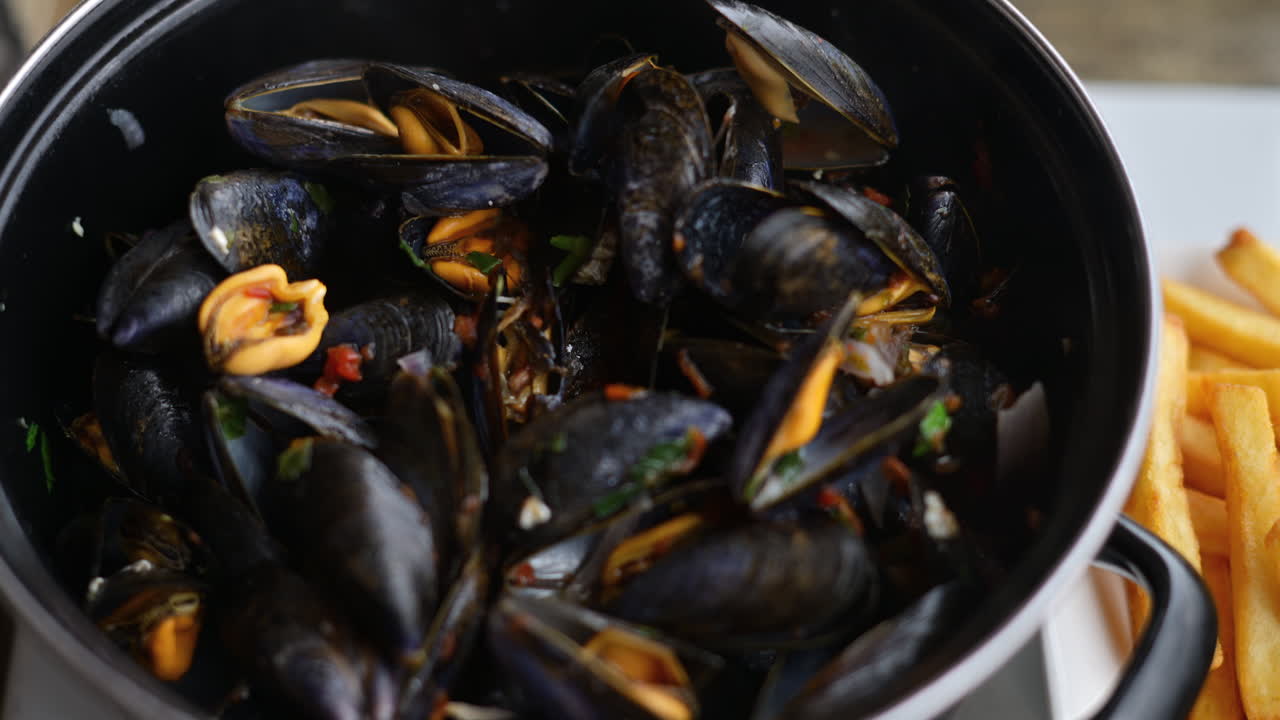 Mejillones cocinados servidos en una olla con hierbas y salsa, acompañados de patatas fritas doradas.