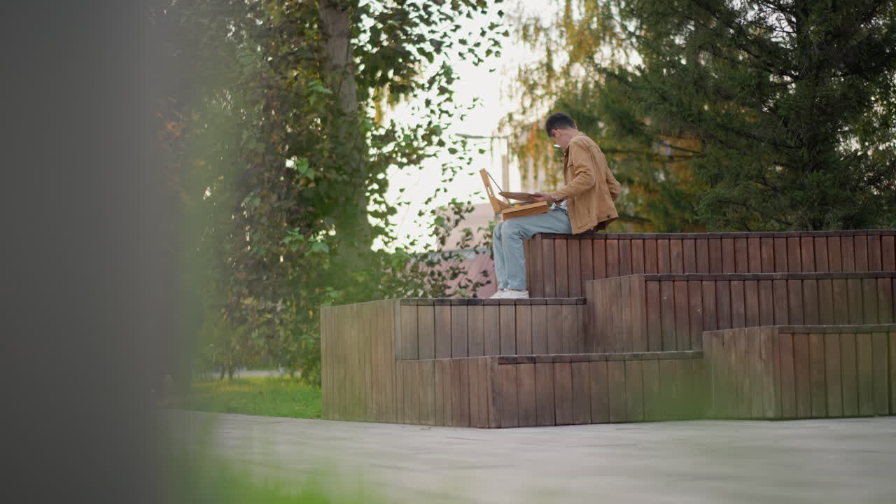 illustrator painting on tiered wooden benches golden hour, sunlight filtering through leaves with watercolor set and brush in hand, wearing jeans and jacket with focused posture, city path