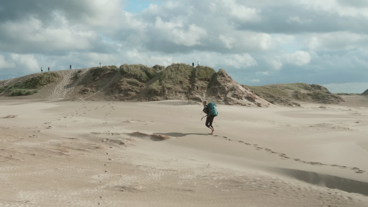 Beautiful Wide Shot of a Lonely Blonde Female Hiker with Heavy Backpack Walking Up Barefoot a Sand Dune in the Desert on a Sunny Summer Day, R&aring;bjerg Mile, Denmark