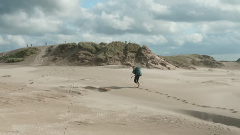 beautiful wide shot of a lonely blonde female hiker with heavy backpack walking up barefoot a sand dune in the desert on a sunny summer day, råbjerg mile, denmark