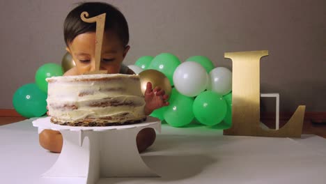lindo bebé latino celebrando su cumpleaños mordiendo su pastel y sonriendo con globos verdes en el fondo