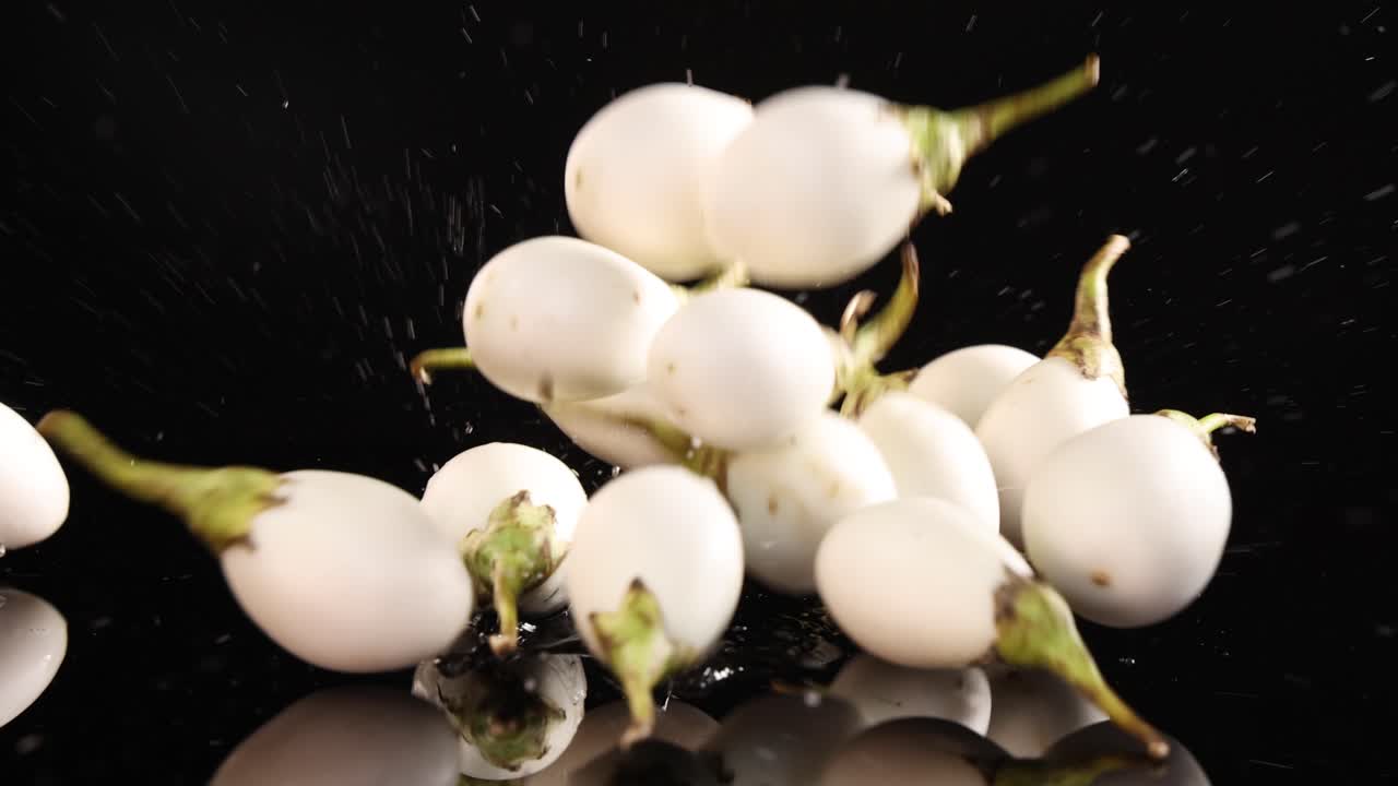 Several white Thai eggplants fall and bounce on a glossy black surface, captured in slow motion with dramatic studio lighting and high contrast