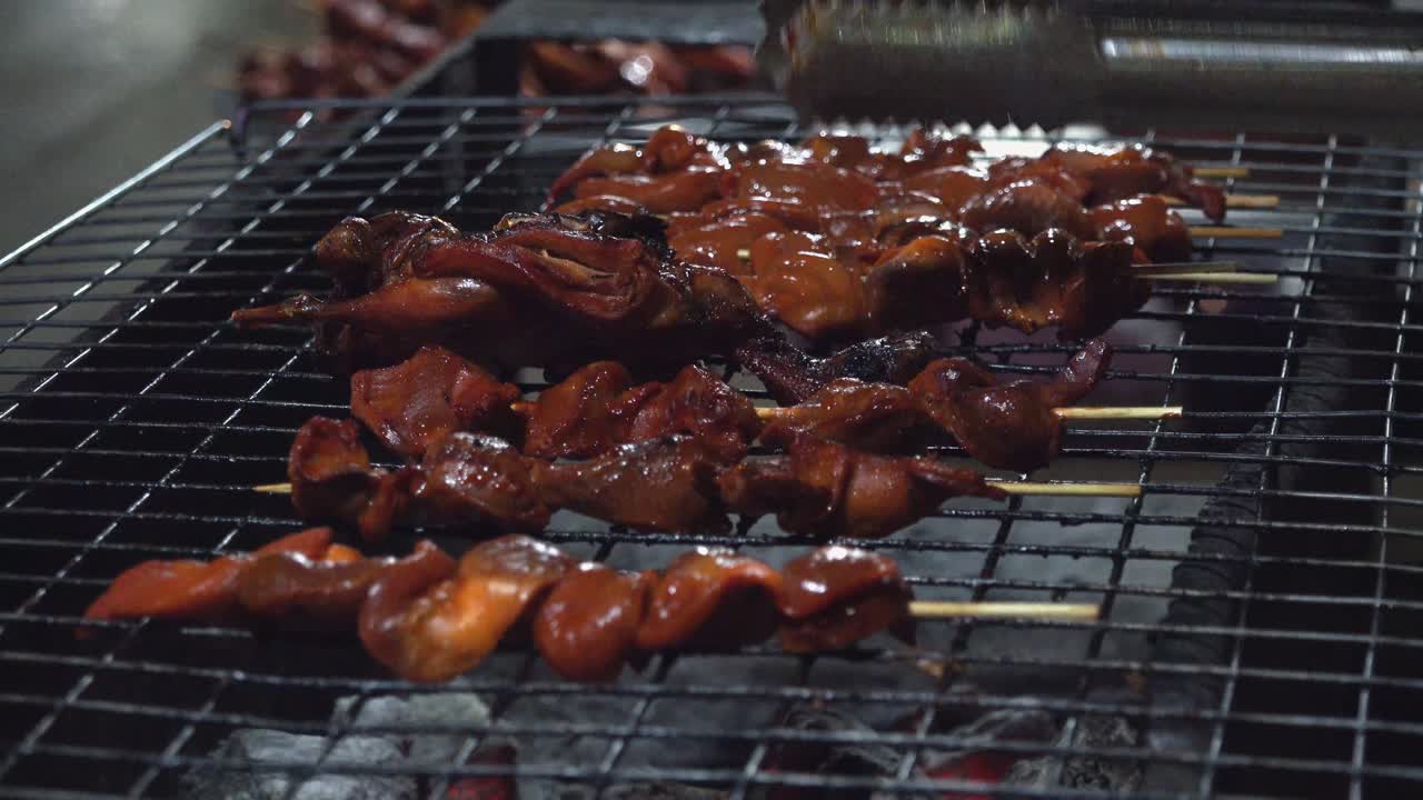 Close Exterior Still shot of Chicken and Pork Insides on a Stick being Flipped Over On a Grill with Tongs at a Street Food Stall In the Night Time
