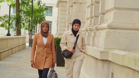 two muslim businesswomen wearing hijabs with modern business suits walking to work past city office buildings 2