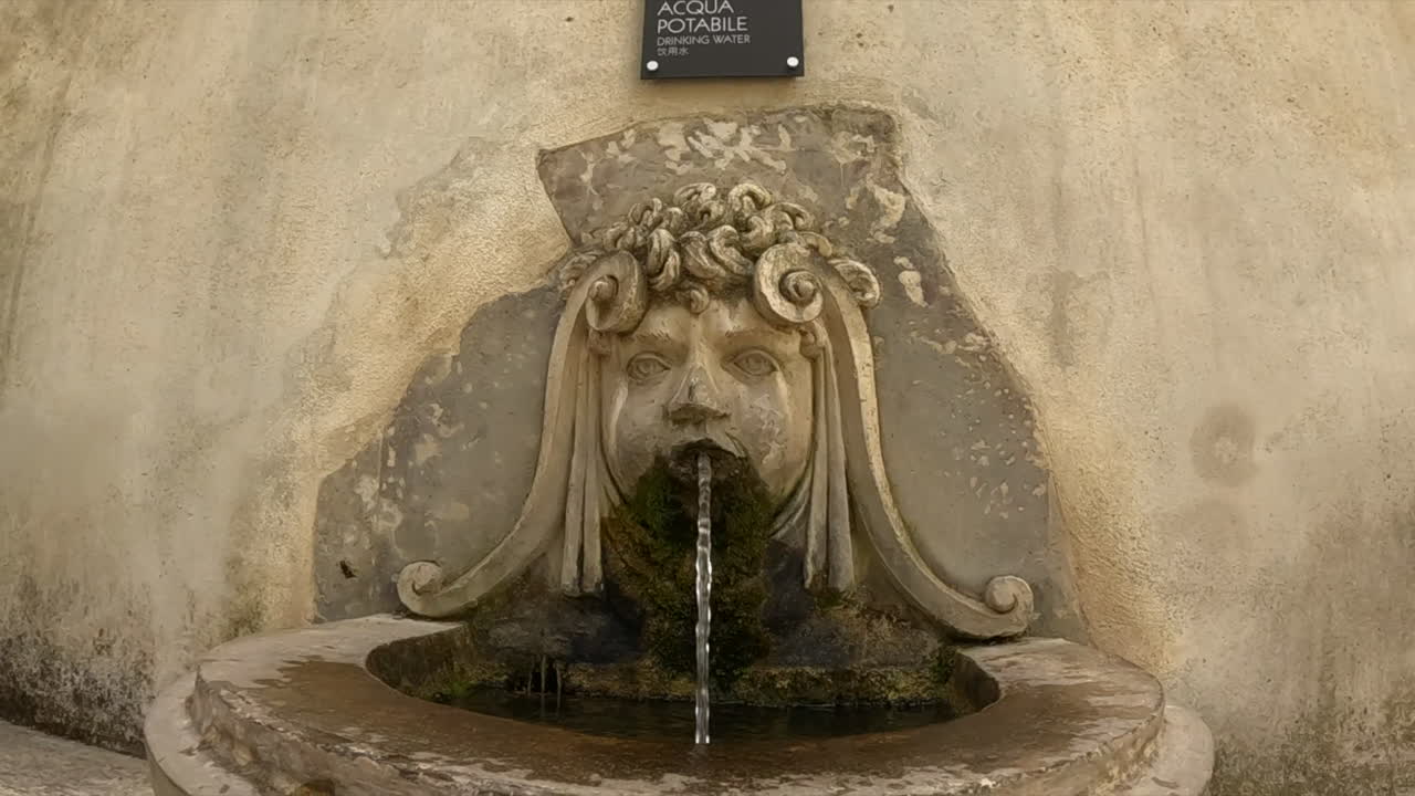 Storica fontana a muro in pietra con una scultura classica di una maschera grottesca che sputa acqua a Tivoli, Roma. Architettura italiana antica con muschio e texture di pietra invecchiata