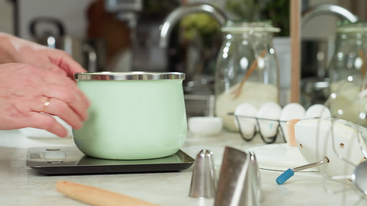 Hand of person places mint green pot on electric stove in bright kitchen setup with eggs, digital scale, colander, glass jar of flour, and utensils on countertop