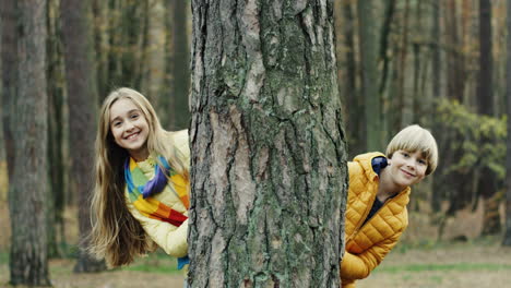 Portrait-shot-of-cheerful-cute-boy-and-girl-looking-at-camera-behind-a-tree-trunk-in-the-forest