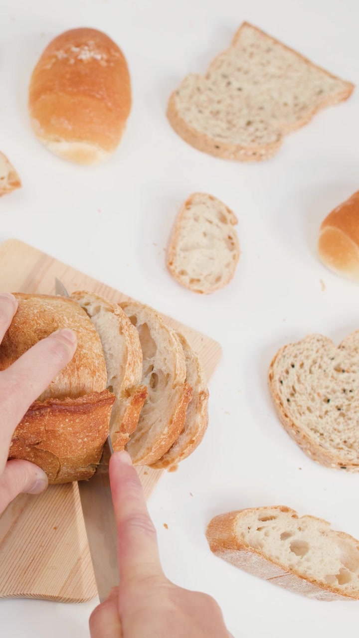 Primo piano dall'alto di mani che tagliano una pagnotta di pane a lievitazione naturale croccante su sfondo bianco con fette di pane