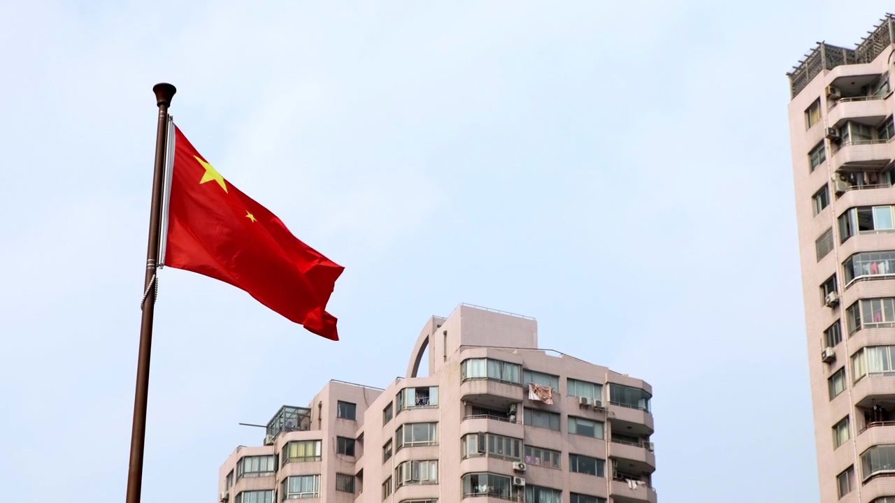 A bandeira nacional da China tremula em um mastro, com edifícios residenciais altos e um céu claro ao fundo.