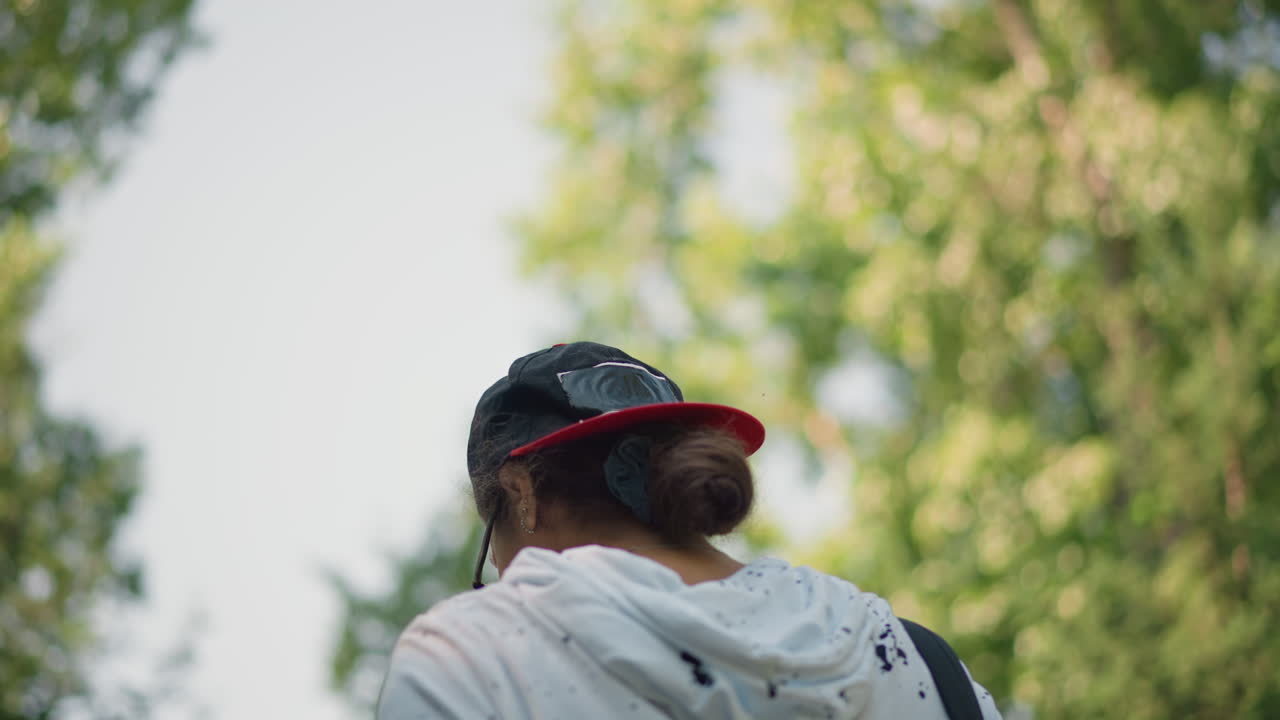 배낭여행객이 모자를 쓰고 후드티를 입고, 머리를 낮게 묶고, 배낭 스트랩이 보이며, 잎이 무성한 덮개가 머리 위에 있고, 부드러운 햇빛이 나뭇잎 사이로 비추고, 느리고 꾸준한 속도로 걷는 뒷모습, 반사되는 야외