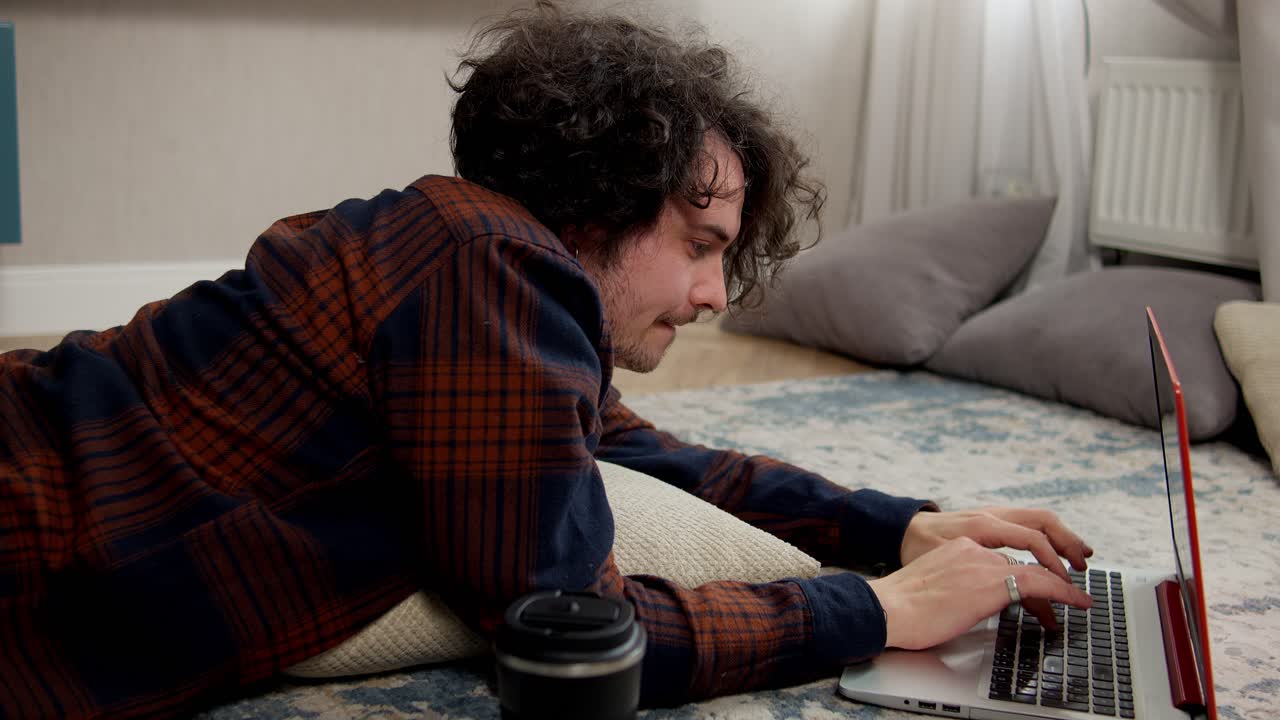 Confident brunette guy with curly hair in a checkered shirt writes his ideas into a laptop while lying on the floor on a pillow in a modern apartment