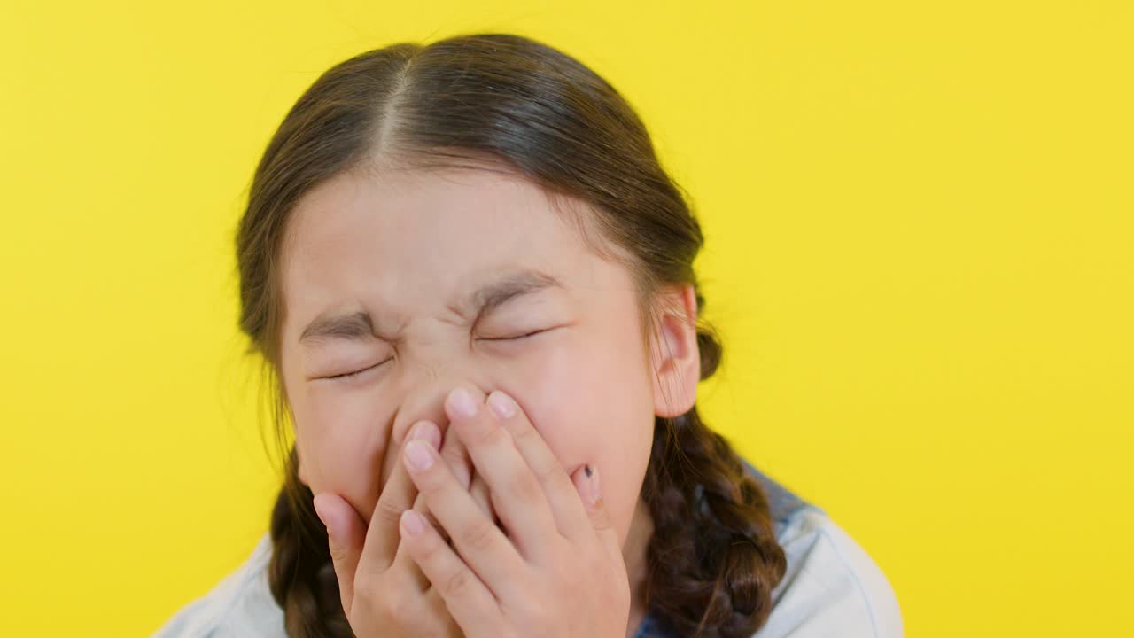 Young girl with braids laughs, covers mouth, and smiles brightly against vibrant yellow backdrop