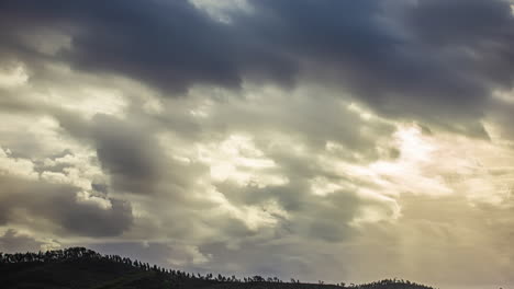 Himmel-Mit-Weißen-Wolken,-Die-Am-Himmel-Vorrücken-Und-Durch-Die-Die-Sonnenstrahlen-über-Einen-Hügel-Mit-Bäumen-Hindurchgehen