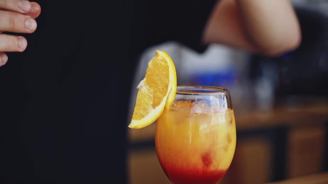 Ready cocktail on a bar table. Bartender puts a slice of orange on the top of the glass with bright beverage. Glass with drink in the nightclub.