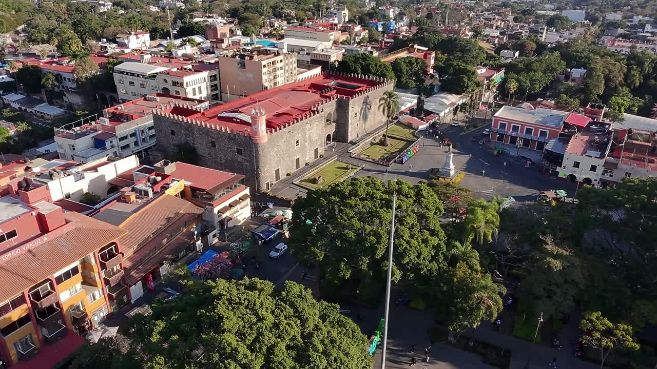 Aerial orbit shot of Cuernavaca city center showing Palacio de Cortés, tree-filled Plaza de Armas, colorful Morelos letters, and the Carlos Pacheco monument under clear afternoon light