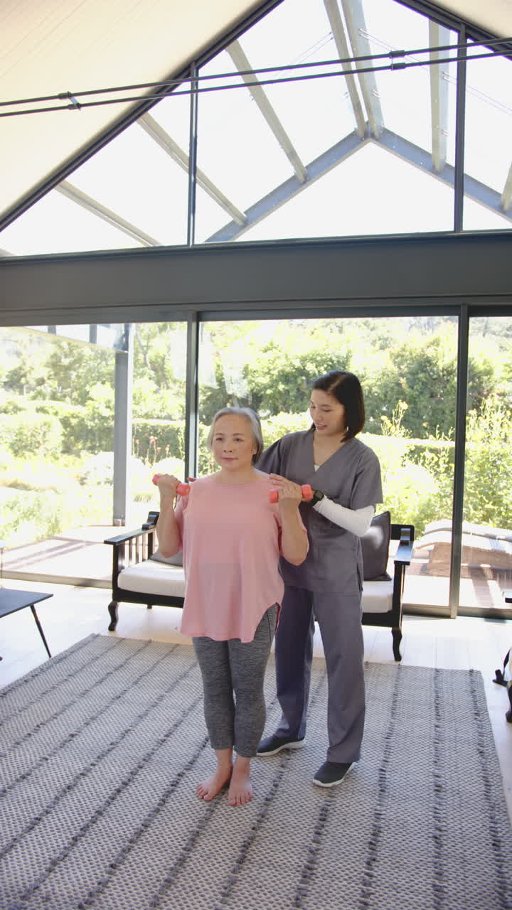 Vertical video: Senior woman exercising with dumbbells, assisted by physiotherapist at home