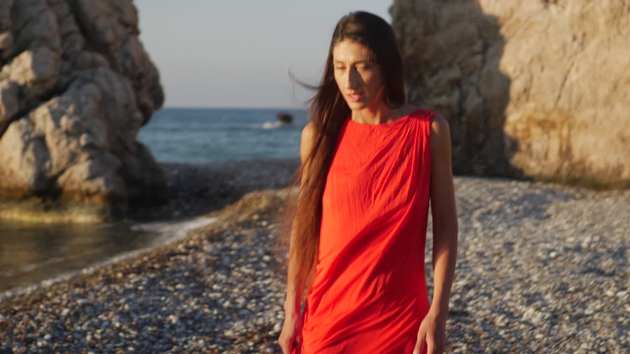 Confident Fashion Female Model in a Red Dress With Long Hair Walking by the Sea on the Beach During Golden Hour, Full Frame