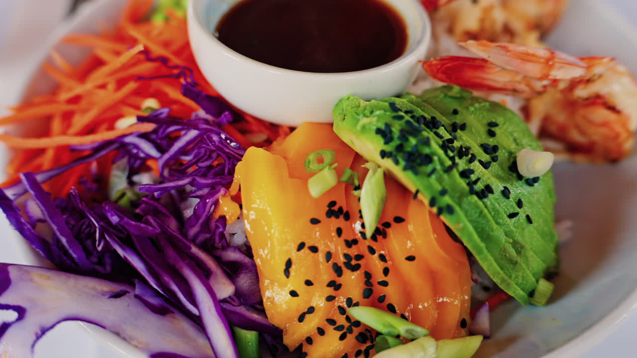 Close up of a poke bowl with rice, avocado, carrot, red cabbage, shrimp and mango and sauce in a white bowl