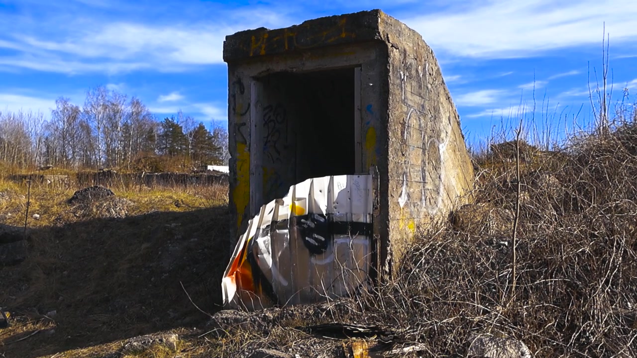 Vecchio ingresso di un bunker militare o rifugio in cemento abbandonato, coperto di graffiti, che si erge in un campo desolato nell'Europa orientale sotto un cielo azzurro e sereno in una giornata di sole.