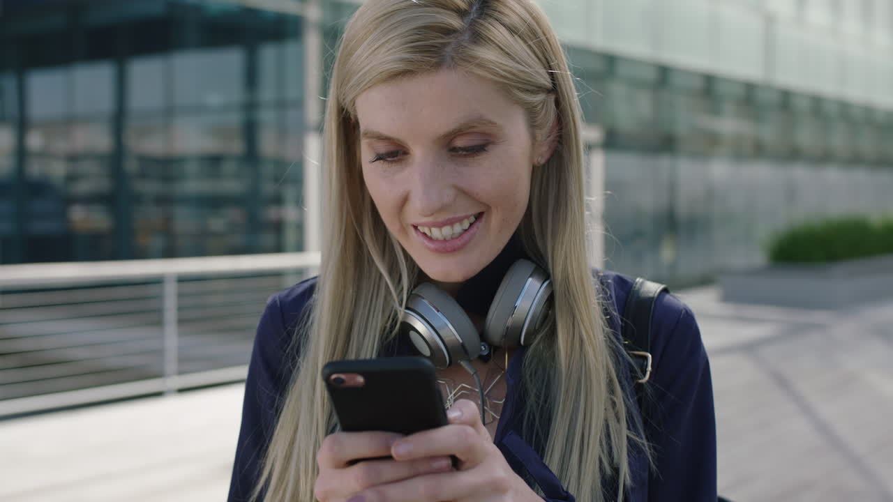 ritratto di una giovane donna d'affari bionda vivace stazionaria sorridente che si gode di messaggi di testo navigando utilizzando l'app di social media dello smartphone in città